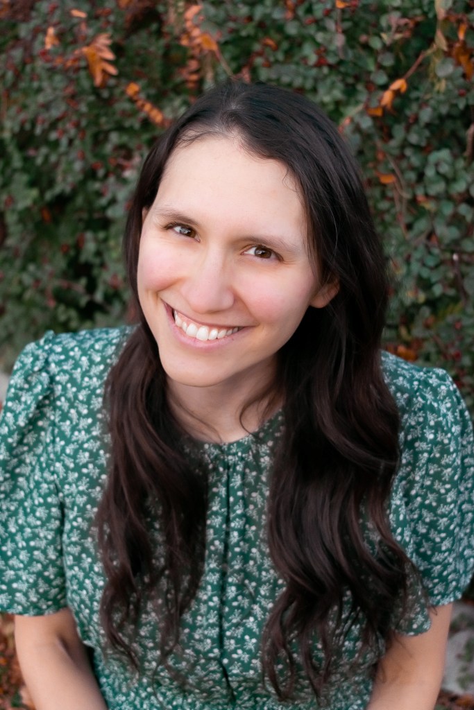 author smiling. brown hair. green floral dress. brown eyes. white skin. bush in background
